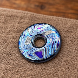 Puede incluir: Un objeto circular y plano con un orificio central, que presenta un patrón arremolinado e iridiscente en tonos metálicos azul, morado y plateado. La superficie muestra una textura compleja y veteada con ondas reflectantes.