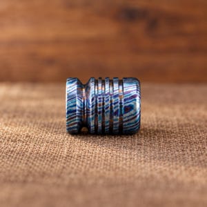 Puede incluir: Una cuenta cilíndrica de metal con un patrón de acero de Damasco multicolor con tonos arremolinados en azul, morado y cobre. La cuenta tiene una serie de ranuras horizontales profundas y paralelas talladas en su superficie exterior.