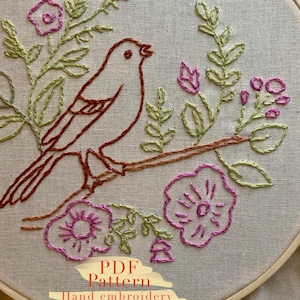 May include: Hand-embroidered bird and floral design in a wooden hoop. The bird is brown, perched on a brown branch, surrounded by green leaves and pink flowers. The text "PDF Pattern Hand embroidery" is at the bottom.