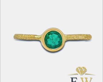 May include: A gold ring with a round, vibrant green emerald gemstone set in a bezel. The band has a textured, hammered appearance. The ring is centered on a light background.