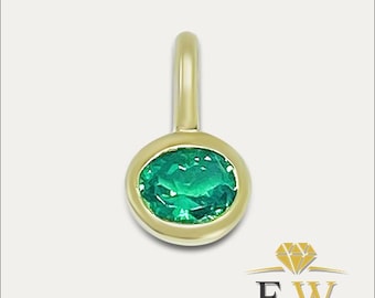 May include: A gold pendant featuring a single oval-cut green emerald gemstone set in a smooth bezel setting. The pendant includes a polished gold bail at the top for attachment to a chain. The design is simple, highlighting the vibrant green stone.