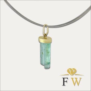 May include: A pendant featuring a raw, light green tourmaline crystal capped with a polished gold bail, suspended from a thin, dark grey wire necklace. The piece displays a natural, elongated mineral structure with a translucent, pale green hue.
