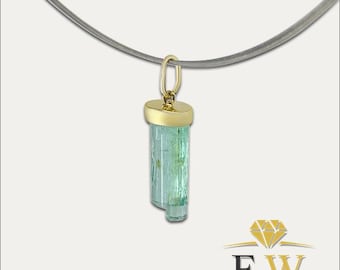 May include: A pendant featuring a raw, light green tourmaline crystal capped with a polished gold bail, suspended from a thin, dark grey wire necklace. The piece displays a natural, elongated mineral structure with a translucent, pale green hue.