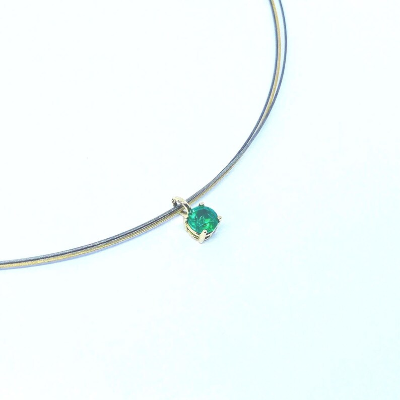 May include: A delicate necklace featuring a vibrant green gemstone pendant. The pendant is set in a gold-colored metal and hangs from a thin, multi-strand chain. The gemstone is round and appears to be emerald. The necklace is displayed against a white background.