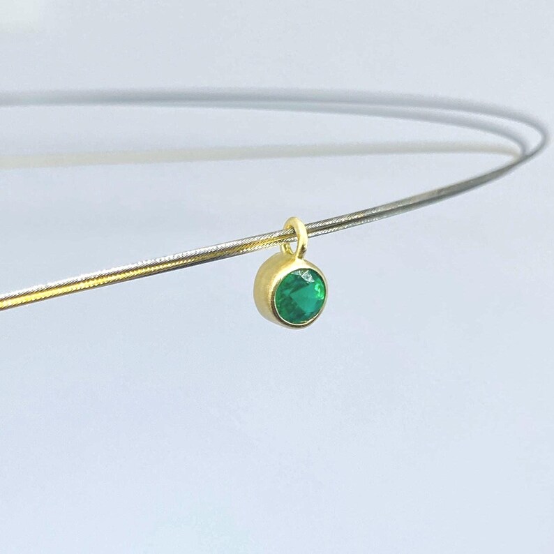 May include: A gold-toned circular pendant featuring a single round, faceted green gemstone set in a bezel. The pendant is suspended on a thin, metallic silver-colored wire necklace that curves across the frame, creating a simple and elegant jewelry piece.