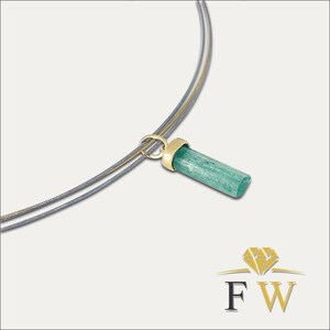 May include: A necklace featuring a cylindrical, light green gemstone pendant capped in polished yellow gold. The pendant hangs from a thin, double-strand grey cord. A logo with a diamond icon and the letters FW is visible in the bottom corner.
