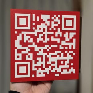 May include: A square, red QR code with a white pattern. The code is held in a person's hand, with the black sleeve of a shirt visible. The background is blurred, with neutral tones.
