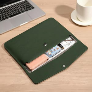 May include: A dark green leather laptop sleeve with a magnetic snap closure. The sleeve is open, revealing a smartphone, glasses, pen, and a small pink case. A laptop and a white mug are in the background.