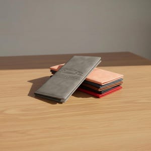 Op de afbeelding: Een stapel portemonnees in diverse kleuren, waaronder grijs, perzik en rood, ligt op een houten oppervlak. De grijze portemonnee op de voorgrond heeft een auto-silhouet en tekst. De portemonnees lijken van leer of een vergelijkbaar materiaal te zijn.