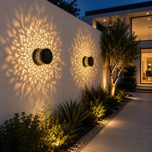 May include: Two circular black solar wall lights mounted on a white exterior wall, casting intricate, large, golden-yellow mandala-shaped light patterns onto the surface. Each fixture features a central solar panel and a perforated metal casing.