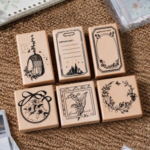 Könnte beinhalten: Ein Set aus sechs rechteckigen Holzstempeln mit schwarzen Tintenmotiven. Die Stempel zeigen Illustrationen eines Hängesessels, einer Liste mit der Aufschrift IMPORTANT, eines Rahmens mit HAPPY DAY CUTE, einer Blume und eines Blumenkranzes.