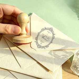 May include: A light-coloured wooden handle stamp with a red base, positioned on a stack of cream-coloured envelopes. One envelope features a black ink oval frame design with the text: Wedding of Sophie & Michael, London 25.07.30.