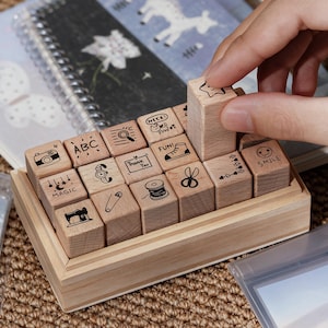 May include: A set of small square wooden stamps arranged in a light wood storage box. Each stamp features a unique black ink icon or text including a camera, ABC, magnifying glass, cat, shoe, scissors, sewing machine, safety pin, spool, and the word SMILE.