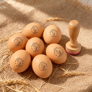 May include: A collection of brown eggs stamped with "JOBELE'S FARM FRESH EGGS" and a hen illustration. A wooden stamp with a pink base sits nearby. The eggs are arranged on a textured, tan surface.