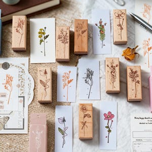 May include: A collection of rectangular wooden stamps featuring various botanical illustrations of flowers and stems, accompanied by several white paper strips displaying stamped floral designs in black, orange, and pink ink colours.