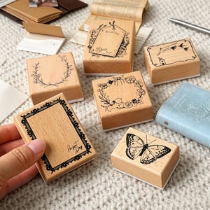 Op de afbeelding: Een verzameling rechthoekige houten rubberen stempels met diverse zwarte inktontwerpen, waaronder een vlinder, een bloemenkrans, een kanten rand met de tekst Perfect Day, een huis met vogels en een labelontwerp met bloemmotieven.