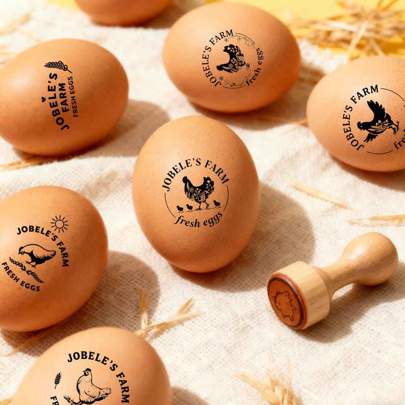 May include: Brown eggs stamped with "JOBELE'S FARM" and "fresh eggs" in black ink, featuring various hen and chick illustrations. A wooden stamp with a hen design is also present. The eggs are on a light-colored surface.