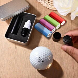 May include: A golf-themed gift set featuring a white golf ball with crossed golf clubs and the initials "BK". The set includes a black ink bottle, a black stamp, and colorful ink pens in a metal case.