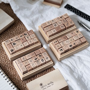 May include: Three rectangular wooden boxes containing sets of small square rubber stamps with various black ink icons and text designs. The stamps feature illustrations of daily items, weather, and activities. A wooden lid labelled DECO desig is included.