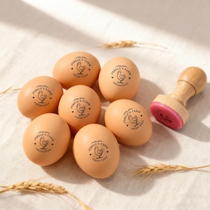 May include: A collection of brown eggs stamped with "JOBELE'S FARM FRESH EGGS" and a chicken graphic. A wooden stamp with a pink ink pad is next to the eggs. The eggs are arranged on a white surface.