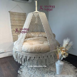 Könnte beinhalten: Ein cremefarbener Makramee-Schaukelstuhl mit Fransenrock und Baldachin. Der Stuhl hat ein weiches, rundes Kissen und ist von der Decke abgehängt. Ein passender Teppich und Wandbehang sind ebenfalls zu sehen.
