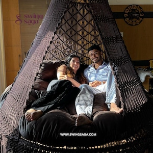 May include: A large, brown macrame swing chair with a fringed bottom and a conical top. The chair has a dark brown cushion and is suspended from the ceiling. The website www.swingsaga.com is visible at the bottom.