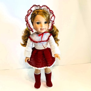 May include: A doll wearing a handmade crochet outfit. The outfit includes a white and red top, a red skirt, a matching bonnet, and red boots. The doll has blonde hair styled in pigtails.