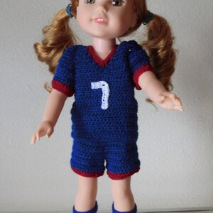 May include: A doll wearing a blue and red crocheted soccer uniform with the number 7 on the front. The doll has long, curly red hair and is wearing white and red crocheted socks.
