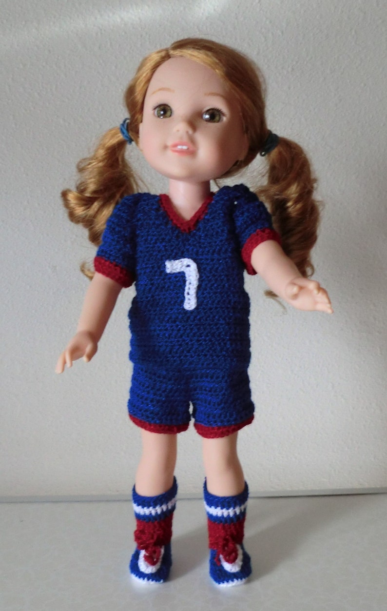 May include: A doll wearing a blue and red crocheted soccer uniform with the number 7 on the front. The doll has long, curly red hair and is wearing white and red crocheted socks.