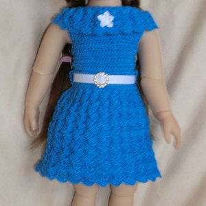 Könnte beinhalten: Eine Puppe mit langen braunen Haaren, die ein blaues Häkelkleid mit einem weißen Blumenakzent und einem weißen Gürtel mit einer goldenen Schnalle trägt. Die Puppe trägt blaue Häkelschuhe.