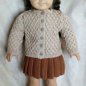 Puede incluir: Una muñeca con un cárdigan de punto beige con un patrón de diamante y una falda y zapatos de ganchillo marrones.