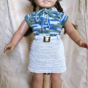 May include: A doll wearing a crocheted striped shirt, a crocheted skirt with a belt, and a straw hat. The doll has brown hair and blue eyes.