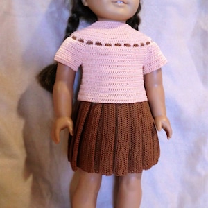 May include: A doll wearing a pink crocheted top with brown trim and a brown crocheted skirt. The doll has brown hair and blue eyes.