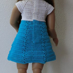 May include: A doll wearing a white crocheted dress with a blue crocheted skirt and a white crocheted hat with a blue ribbon.