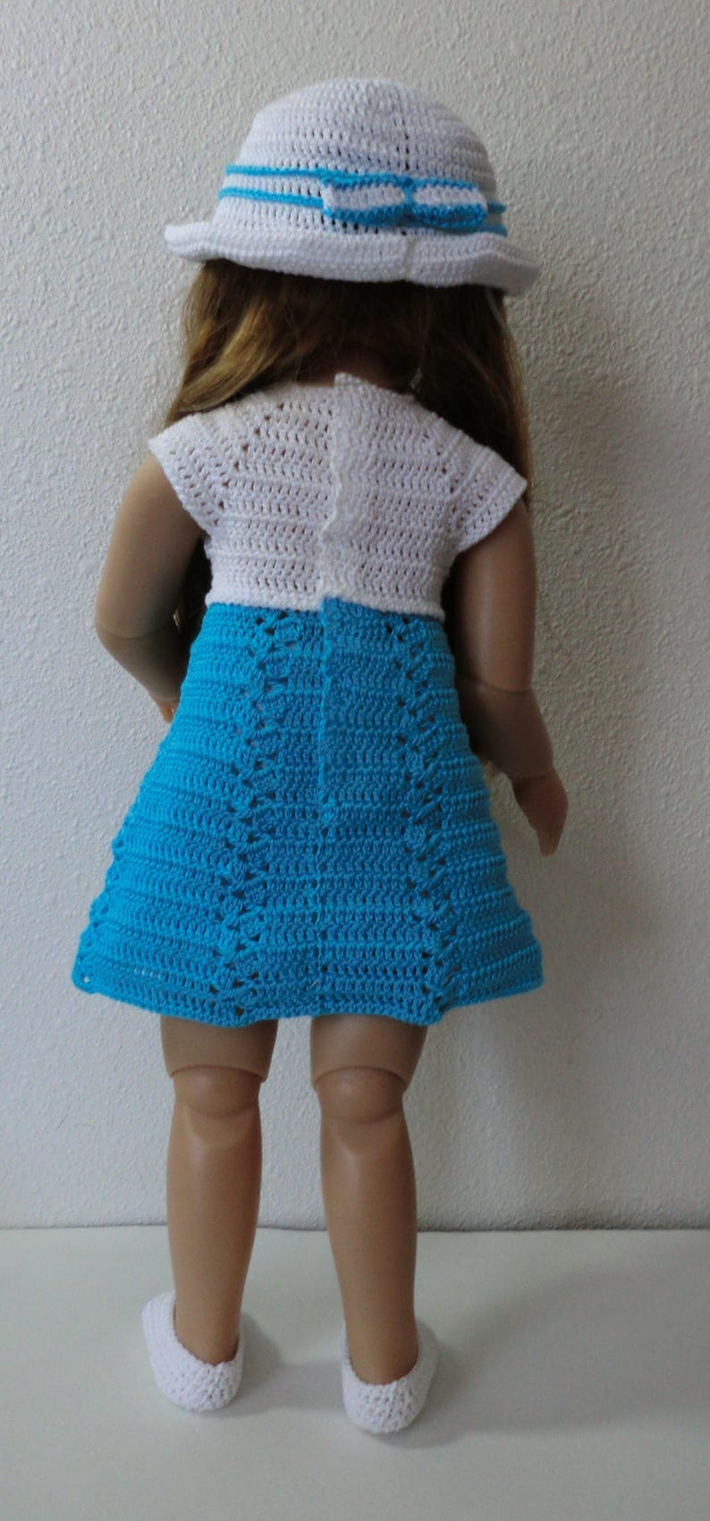 May include: A doll wearing a white crocheted dress with a blue crocheted skirt and a white crocheted hat with a blue ribbon.