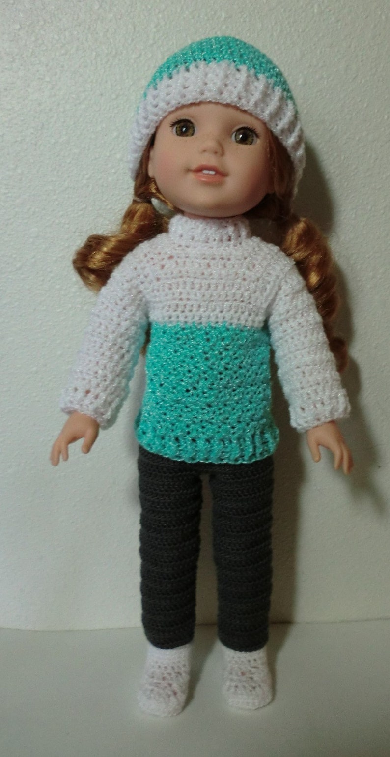 May include: A doll wearing a white and turquoise crocheted sweater, gray crocheted pants, and a white crocheted hat with a turquoise band.