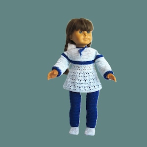 Puede incluir: Muñeca vestida con un conjunto de ganchillo hecho a mano. El conjunto incluye una túnica blanca y azul con mangas largas, combinada con pantalones azules y zapatos blancos. La muñeca tiene el pelo castaño.