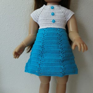 May include: A doll wearing a white crocheted hat, a white crocheted top with blue buttons, and a blue crocheted skirt. The doll is wearing white crocheted shoes.