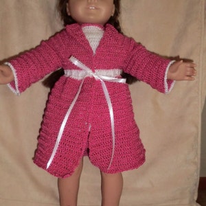 May include: A doll wearing a pink crocheted robe with a white sash and white crocheted slippers.