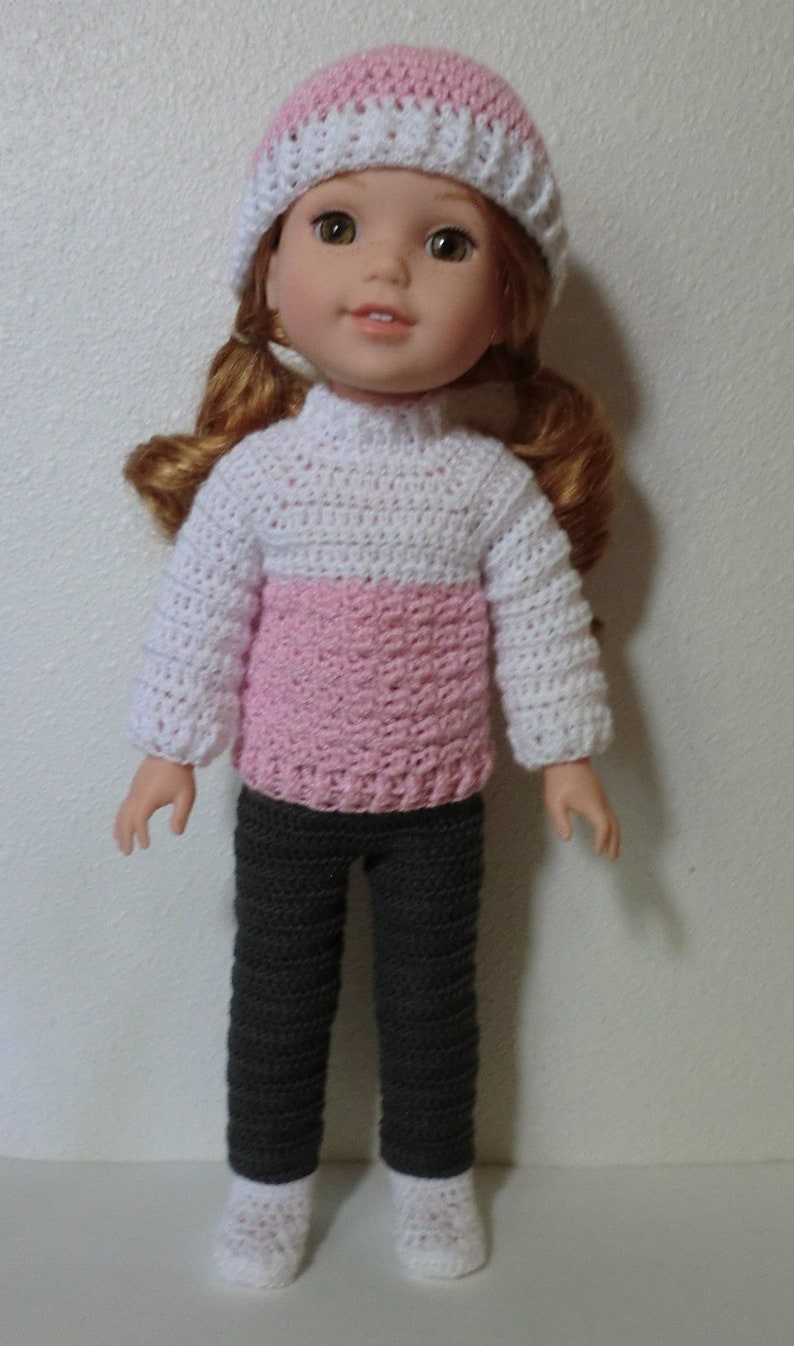 May include: A doll wearing a pink and white crocheted hat, a pink and white crocheted sweater, gray crocheted pants, and white crocheted socks.