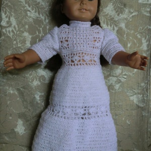 May include: A white crocheted dress with a lacy design. The dress has a high neck and long sleeves. The dress is worn by a doll with brown hair.