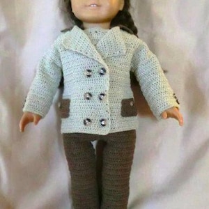 Puede incluir: Una muñeca con una chaqueta de punto de color verde claro con pantalones de punto marrón y cuatro botones. La muñeca tiene el pelo castaño y los ojos azules.