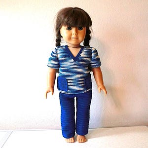 May include: A doll with brown hair in two braids wearing a short-sleeved, V-neck crochet top featuring a variegated blue and white striped pattern with two front pockets, paired with solid dark blue crochet pants that reach the ankles.