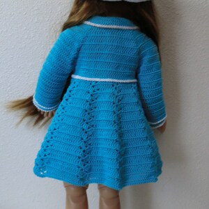 May include: A doll wearing a blue crocheted dress with white trim and a white crocheted hat with a blue ribbon. The doll is wearing white crocheted shoes.