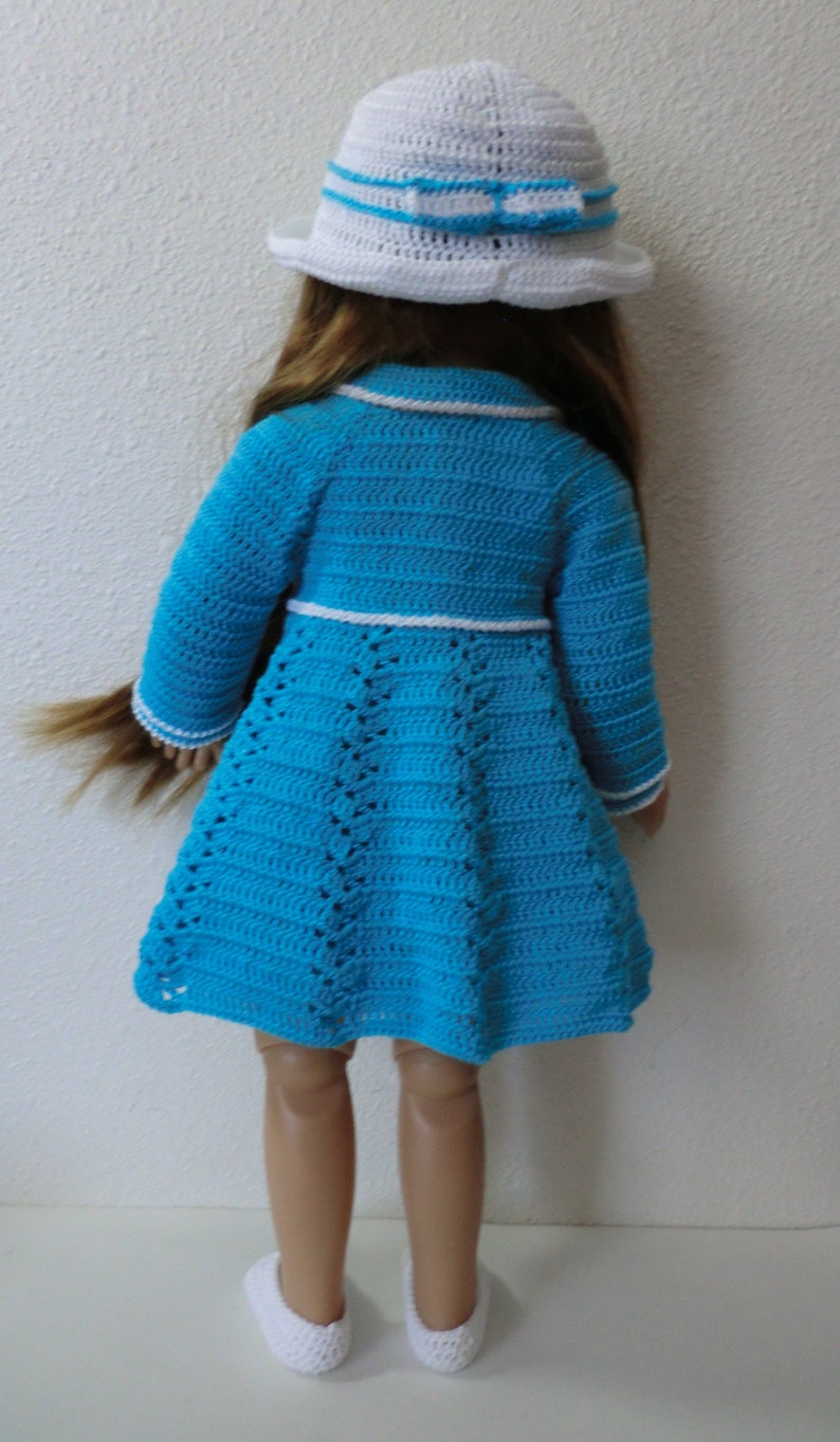 May include: A doll wearing a blue crocheted dress with white trim and a white crocheted hat with a blue ribbon. The doll is wearing white crocheted shoes.