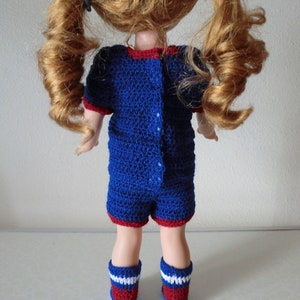 May include: A blue and red crocheted doll outfit with a button closure in the back. The outfit is worn by a doll with long, curly red hair.