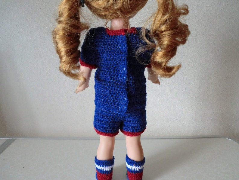 May include: A blue and red crocheted doll outfit with a button closure in the back. The outfit is worn by a doll with long, curly red hair.