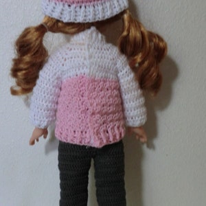 May include: A doll wearing a pink and white crocheted sweater, a pink crocheted hat, and gray crocheted pants.