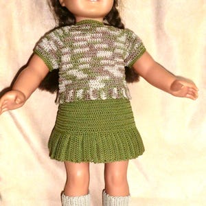 Puede incluir: Una muñeca con un vestido de ganchillo verde y blanco y zapatos de ganchillo marrones. La muñeca tiene el pelo castaño y los ojos azules.