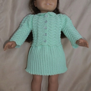May include: A light green knitted dress and cardigan set for a doll. The dress has a skirt with a pleated design and the cardigan has a button closure.